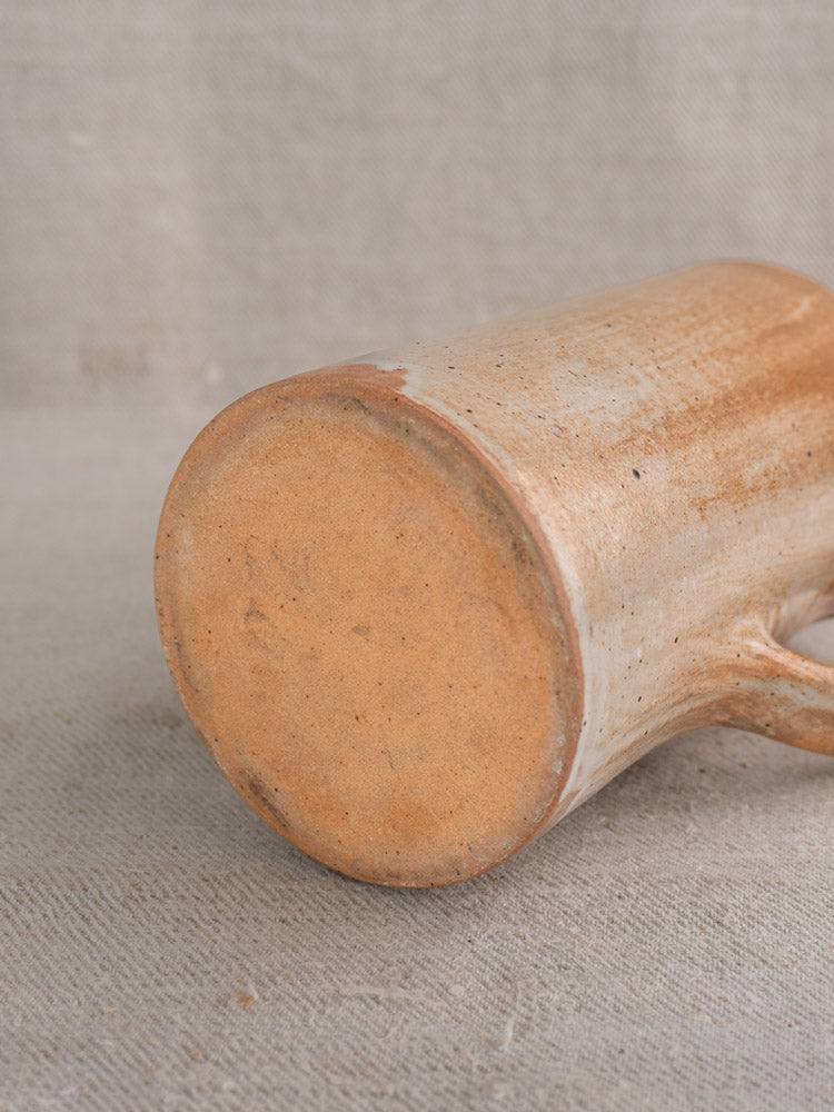 Antique French stoneware cruche with salt glaze, textured base exposed, on neutral coarse fabric background.