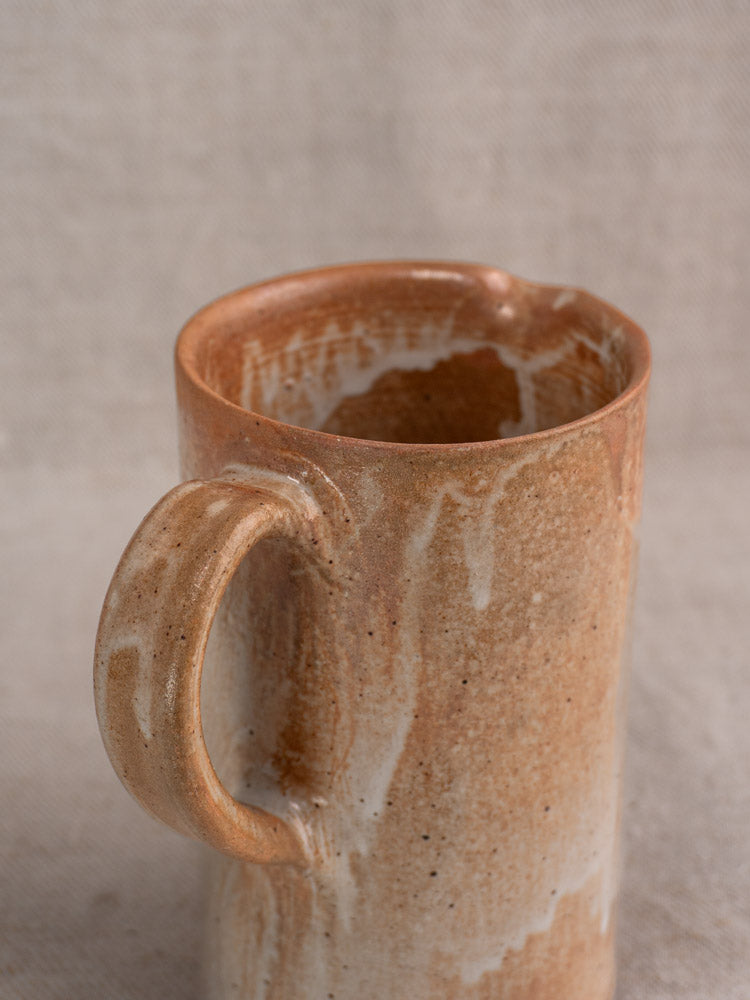 Antique French stoneware cruche with large handle, speckled beige-brown salt glaze, on neutral fabric background.