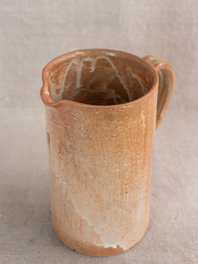 Antique French stoneware cruche with handle, rustic brown salt glaze, and white streaks on a beige textured surface.