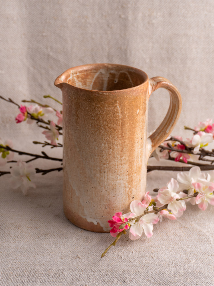 Mid-20th century French stoneware cruche with salt glaze on beige textured fabric, pink and white cherry blossom branches nearby.