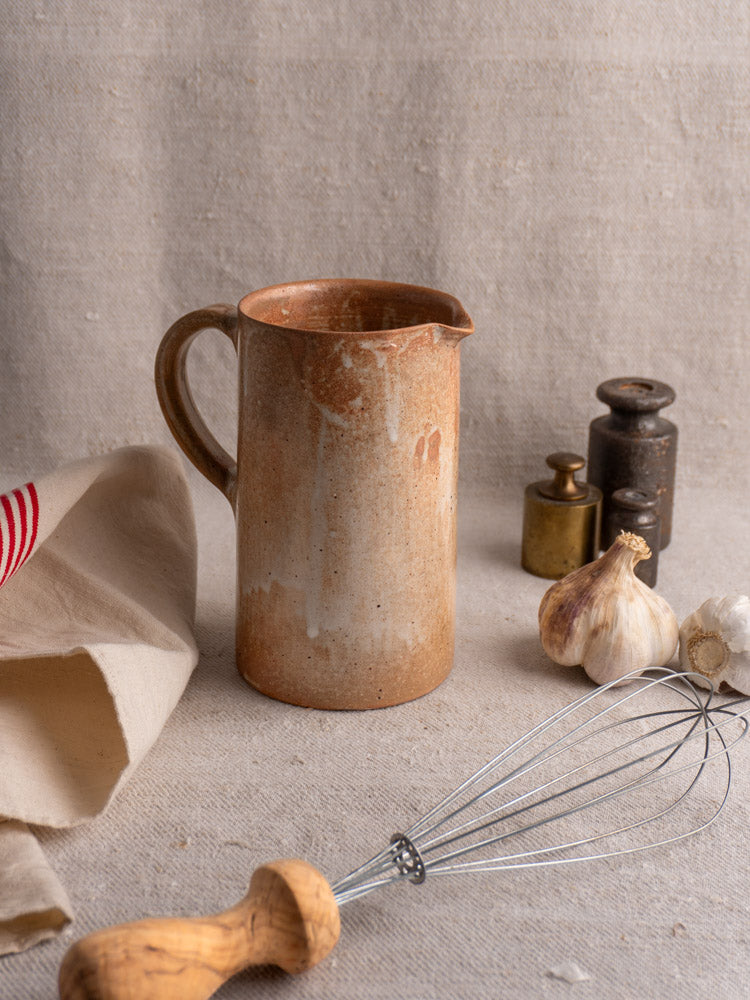 Antique French stoneware pitcher with salt glaze displayed on beige fabric beside garlic bulbs and vintage kitchenware.