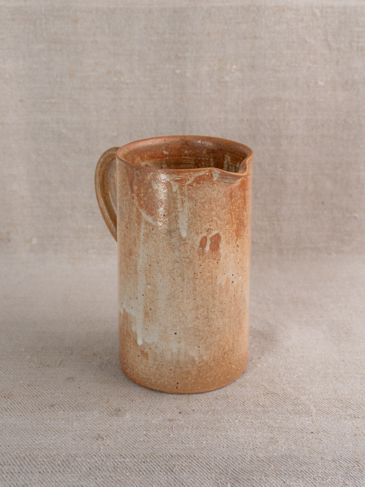 Antique French stoneware cruche with handle and spout in brown and beige glaze on textured beige fabric.