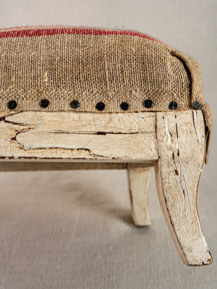 Antique French footstool with Louis XV frame, chipped white paint, hemp torchon upholstery, and exposed tacks.