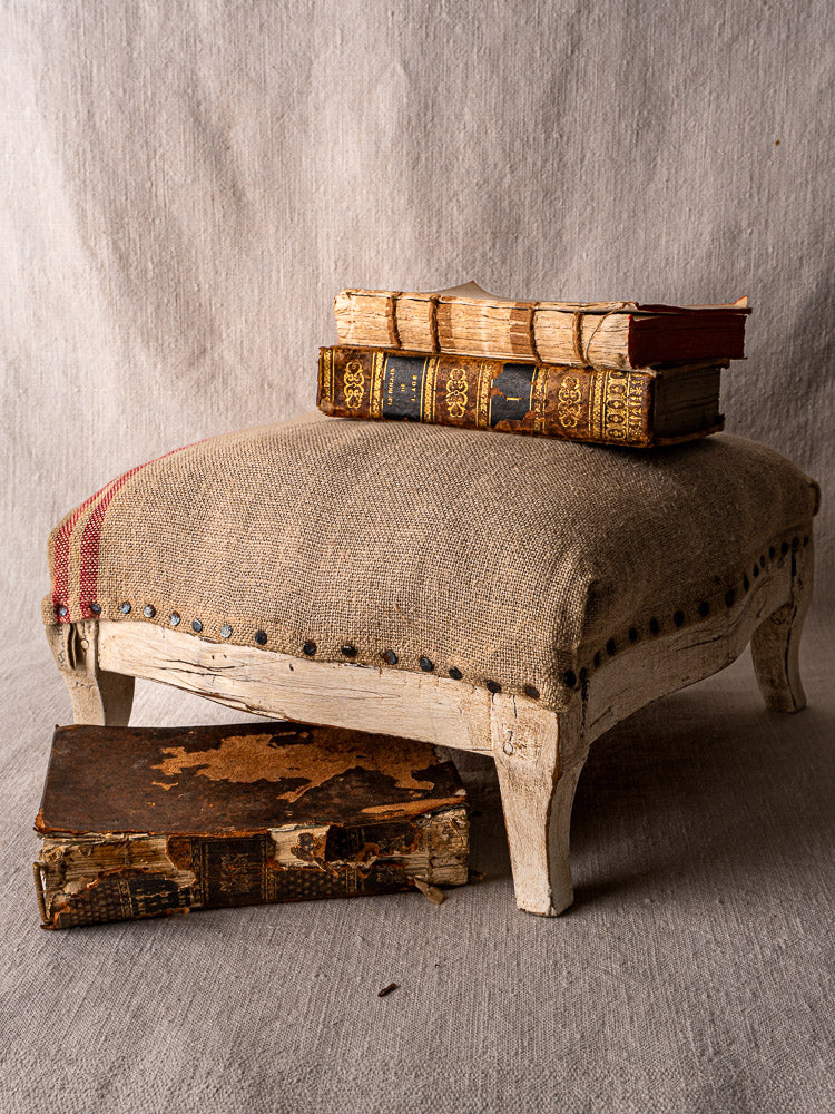 Antique French Louis XV footstool with nailhead trim and rustic hemp upholstery, stacked with old books.
