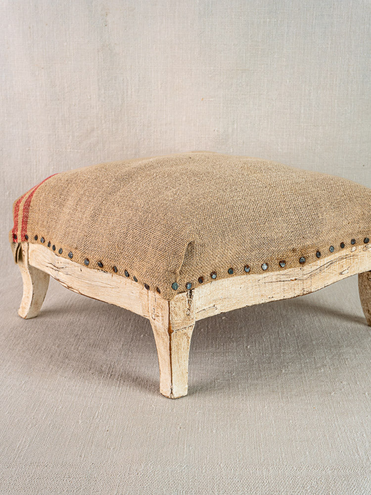 Antique French Louis XV style footstool with painted wood frame, hemp upholstery, burlap accents, and red stripe.