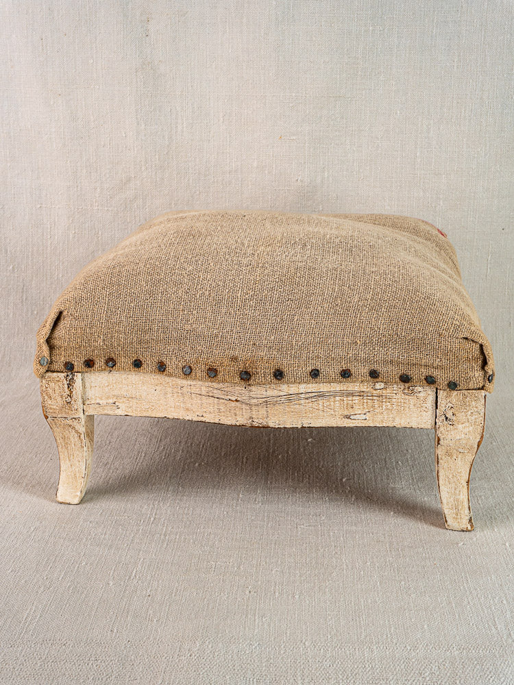 Antique French footstool with painted Louis XV frame, curved legs, hemp torchon seat, on neutral fabric background.