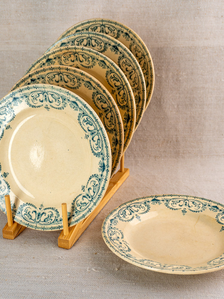 Set of six Antique French Terre de Fer Faience Caïffa Guirlande dinner plates in wooden rack, neutral fabric background.