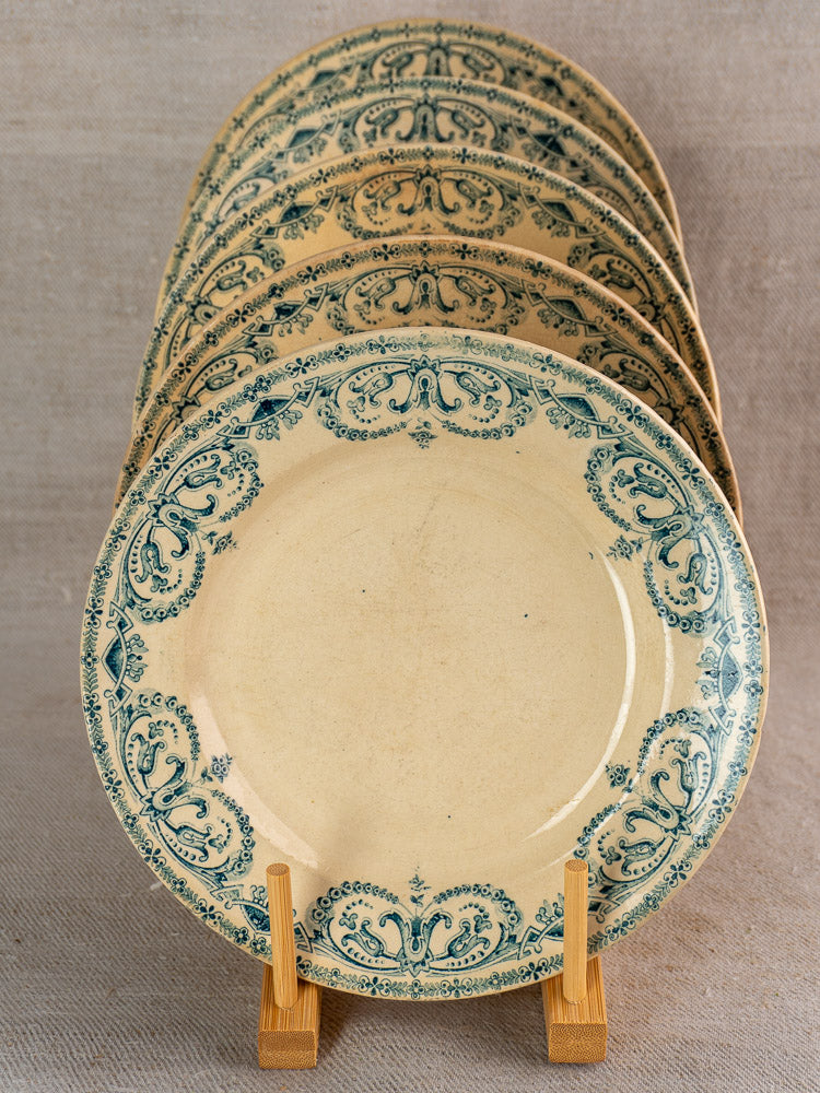 Set of six antique French Terre de Fer faience dinner plates with blue Caïffa Guirlande rims on wooden stand, neutral fabric backdrop.