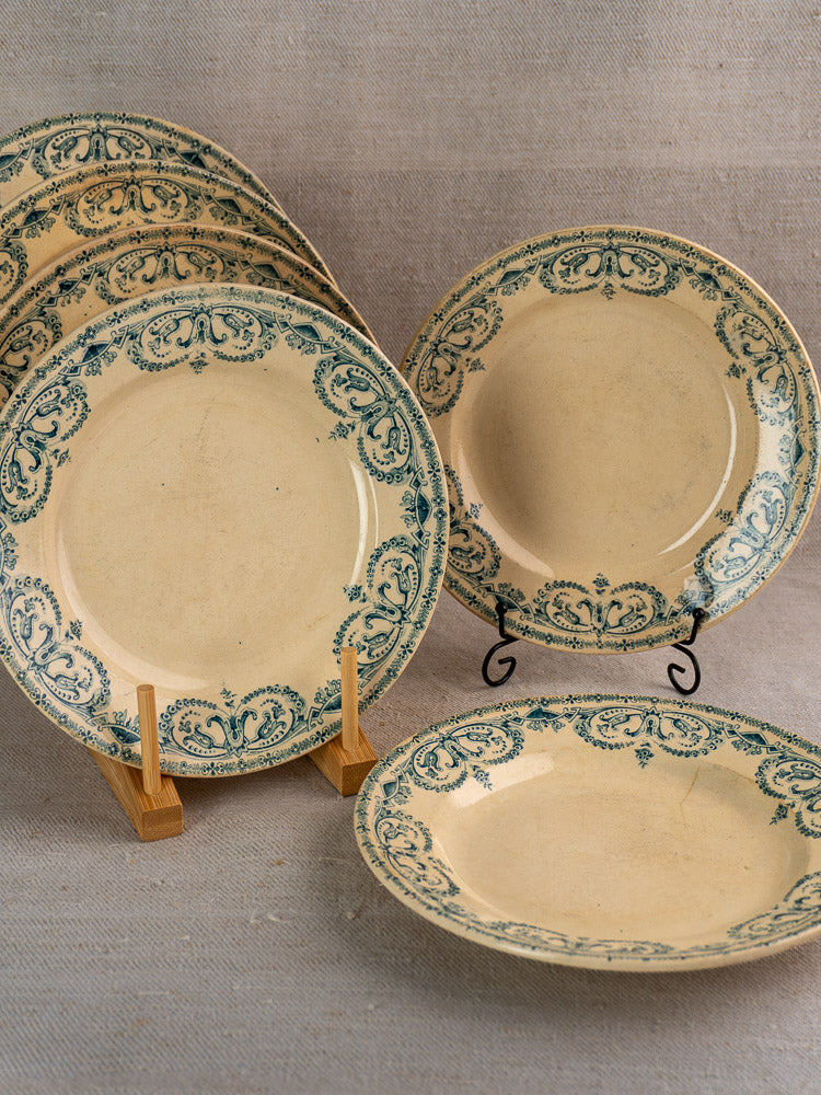 Set of six antique French Terre de Fer faience plates with green Guirlande border, displayed on textured fabric.