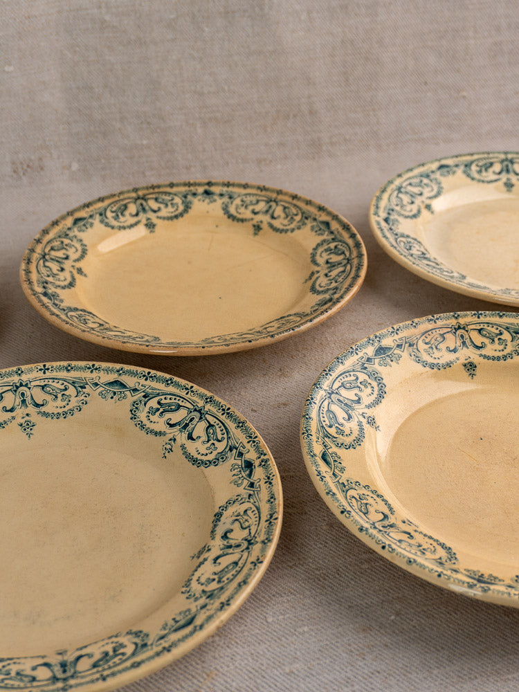 Set of six antique French faience dinner plates with green rim patterns displayed on textured beige fabric.