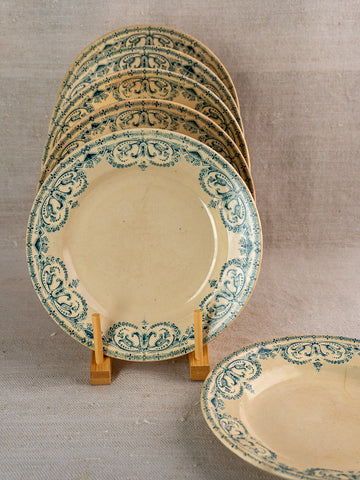 Set of six cream-colored antique French Terre de Fer faience dinner plates on wooden stand, neutral fabric backdrop.