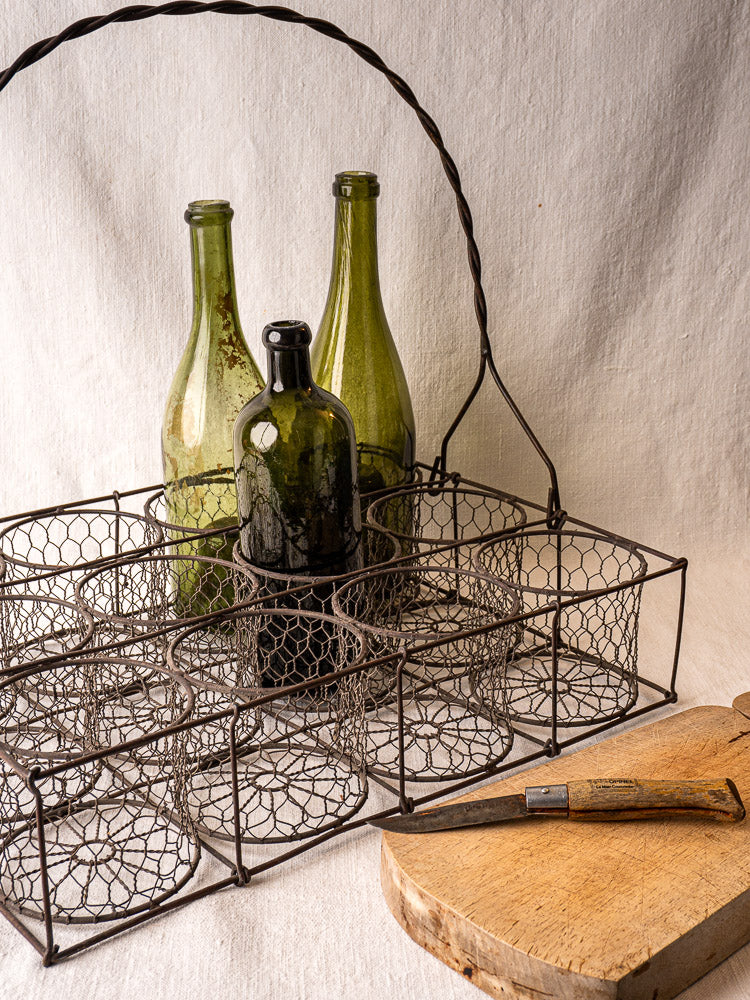 Antique French wire wine caddy with three vintage glass bottles, wooden board, and old knife on light fabric.