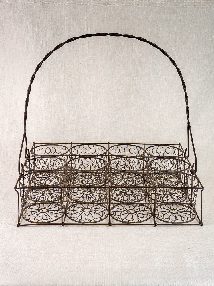 Antique French wire 12-bottle cellar caddy with twisted handle on a light fabric background.
