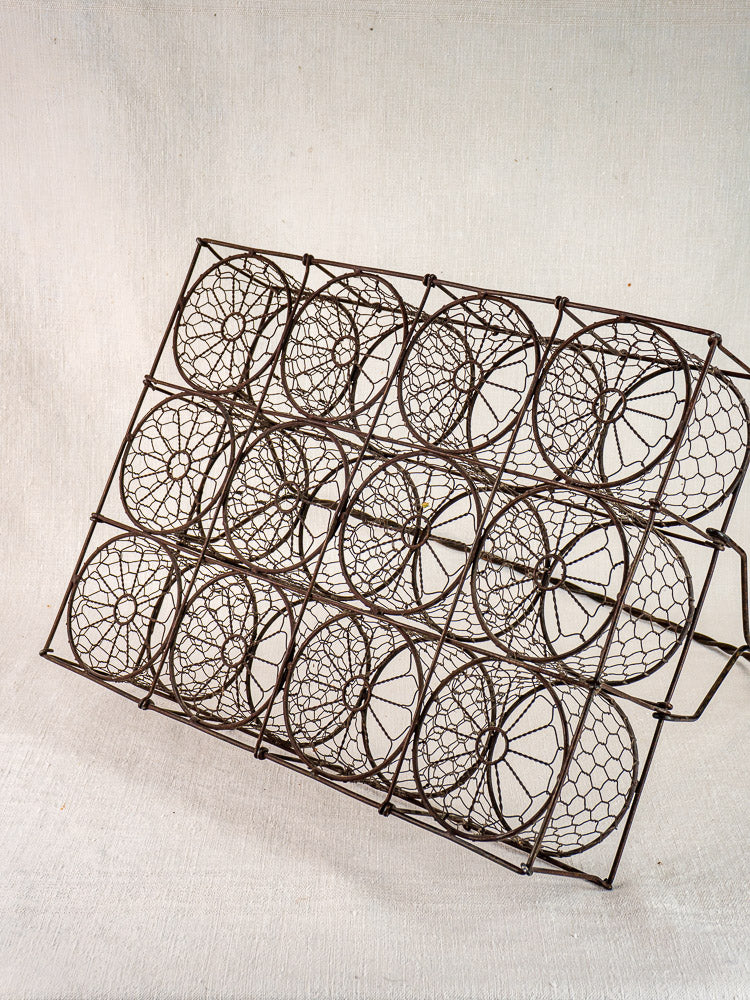 Antique French wire wine carrier with twelve circular holders, angled on a neutral fabric background.