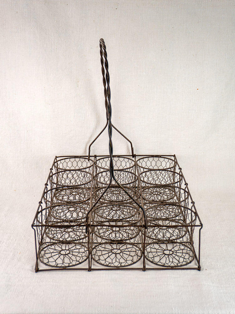 Antique French wire wine carrier with twisted handle and twelve compartments, on neutral fabric background.