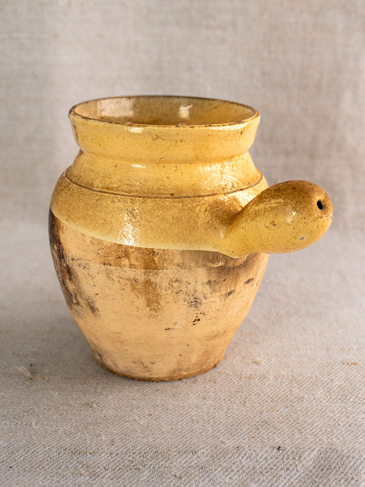 19th-century French terracotta toupin with rustic yellow glaze and round handle, displayed on textured beige fabric.