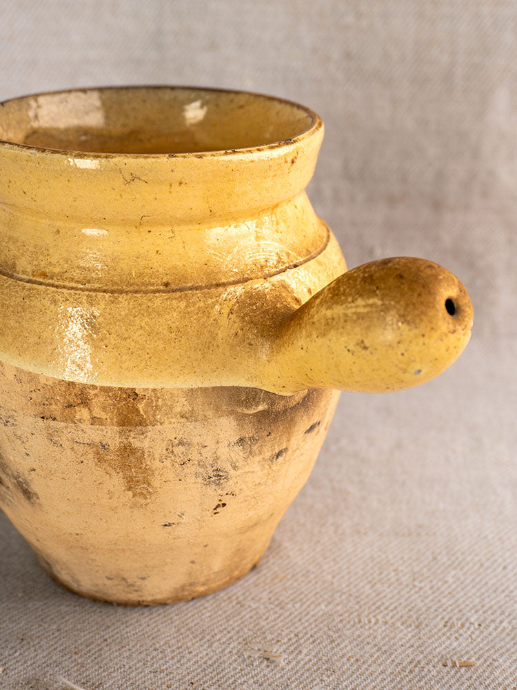 Antique French glazed terracotta toupin with spout handle, rustic yellow finish, on textured beige fabric background.