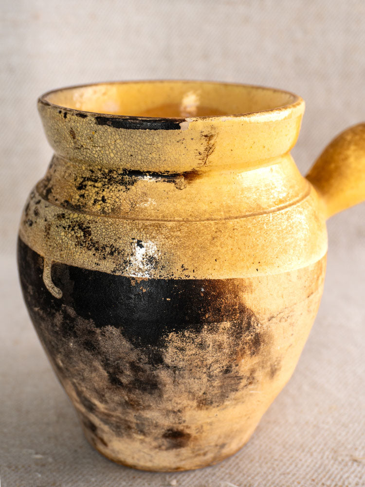 Antique French toupin, glazed terracotta jug with yellow glaze and worn black patches on neutral fabric background.