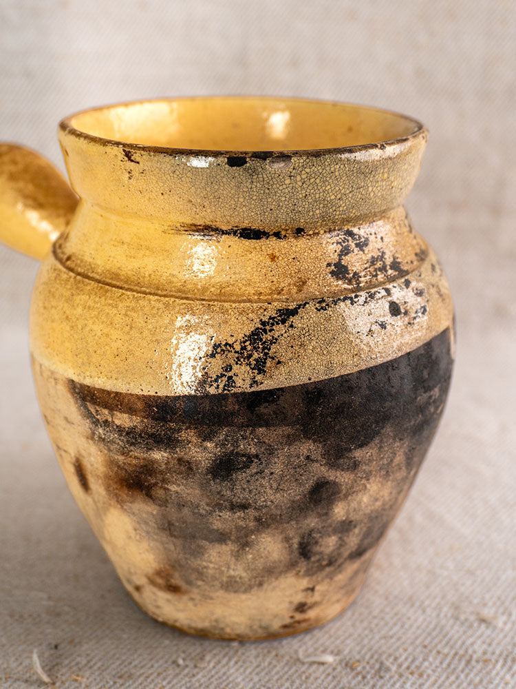 Antique French glazed terracotta toupin with handle, heavy scorch marks, weathered surface on beige textured fabric.
