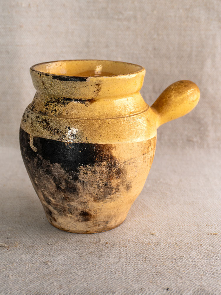 Antique French 19th-century glazed terracotta toupin with single handle and Provençal yellow and black design on beige fabric.
