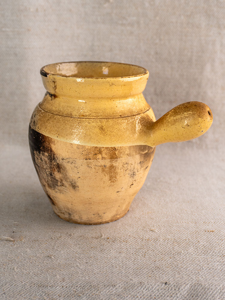 Antique French Toupin, yellowish-brown glazed terracotta with rounded body and sturdy handle on neutral textured fabric background.