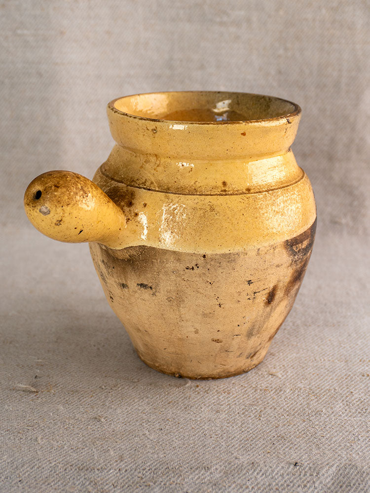 Antique French toupin with rustic yellow glaze, wide mouth, spout handle, and patina on textured grey fabric.