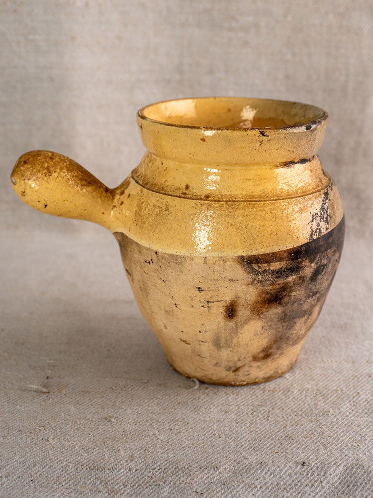 19th-century French toupin with rustic yellow glaze, single handle, rough texture, and burn marks on neutral fabric.