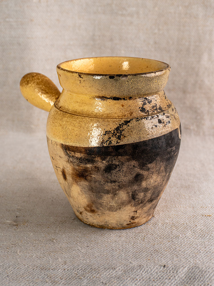 Antique French glazed terracotta toupin with single handle and yellow-brown glaze on textured beige fabric background.