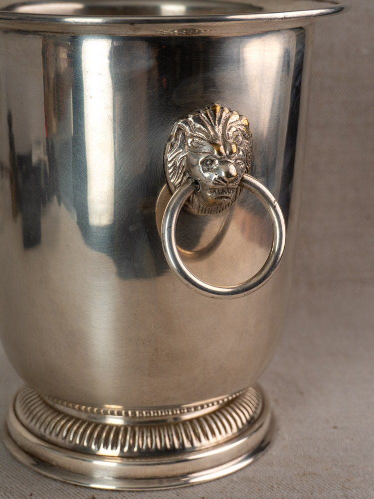 Vintage French silver-plated champagne bucket with lion head handles and ribbed base on a neutral fabric background.