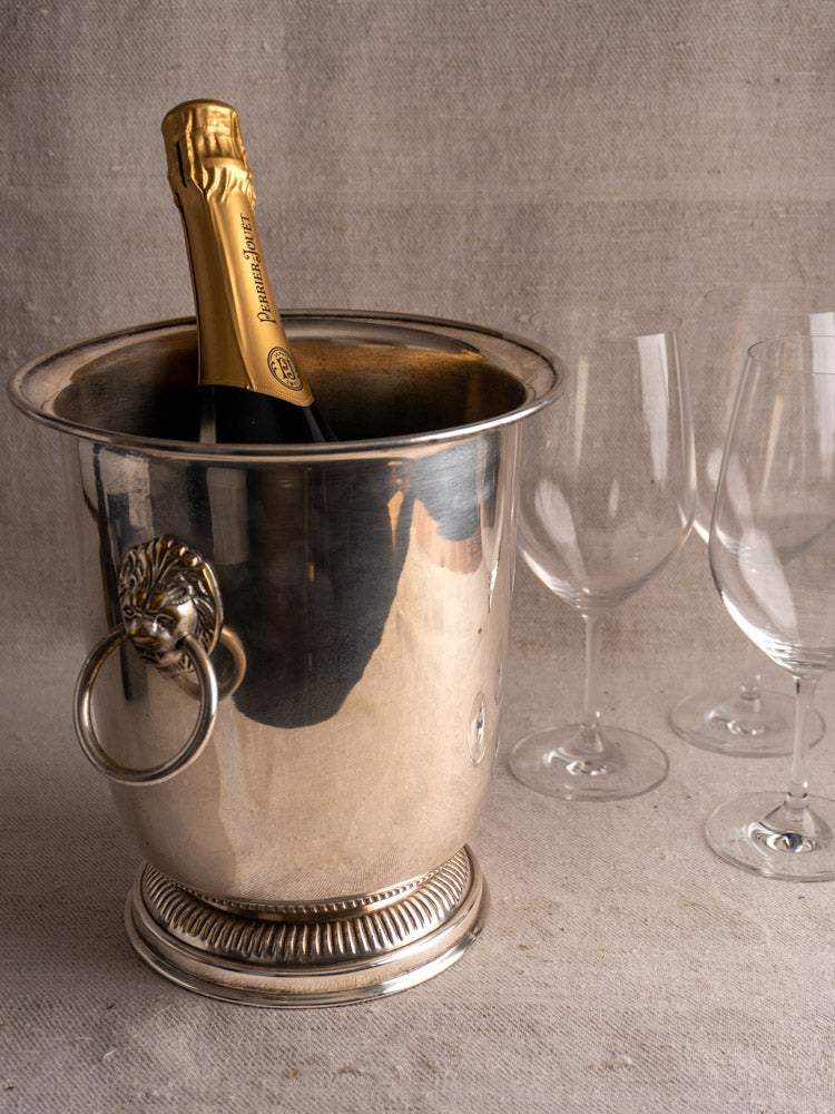 Vintage French silver-plated champagne bucket with lion head handles beside three empty wine glasses on a beige textured surface.