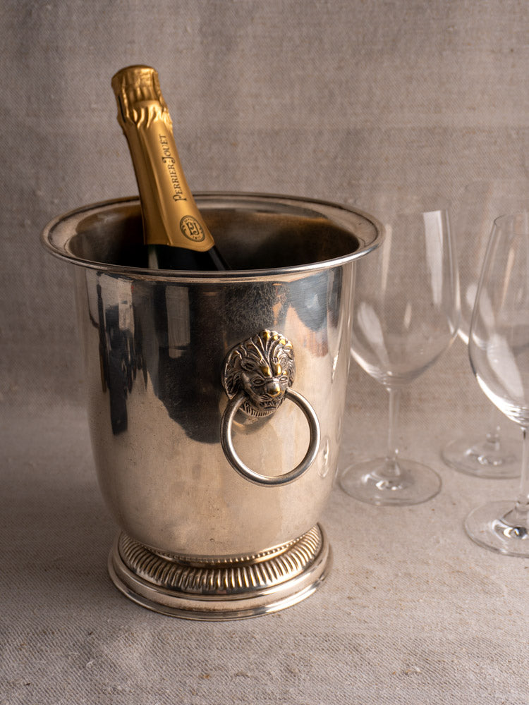 Silver plated vintage French champagne bucket with lion head handles, chilling champagne beside three empty glasses on fabric.