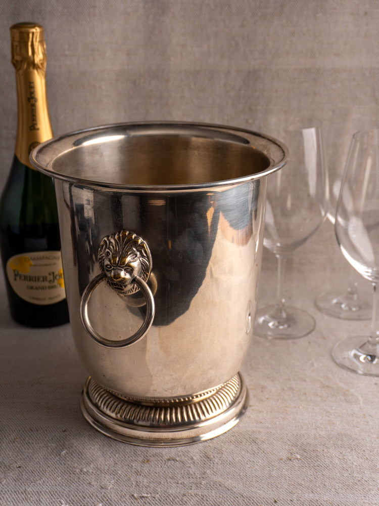 Silver plated vintage French champagne bucket with lion head handles and decorative rings, set on textured beige fabric.
