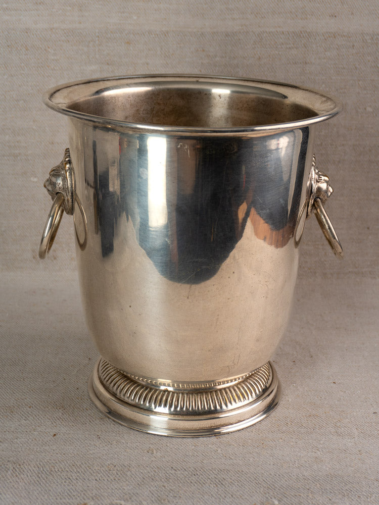 Silver plated vintage French champagne bucket with lion head handles and decorative rings, set on textured beige fabric.