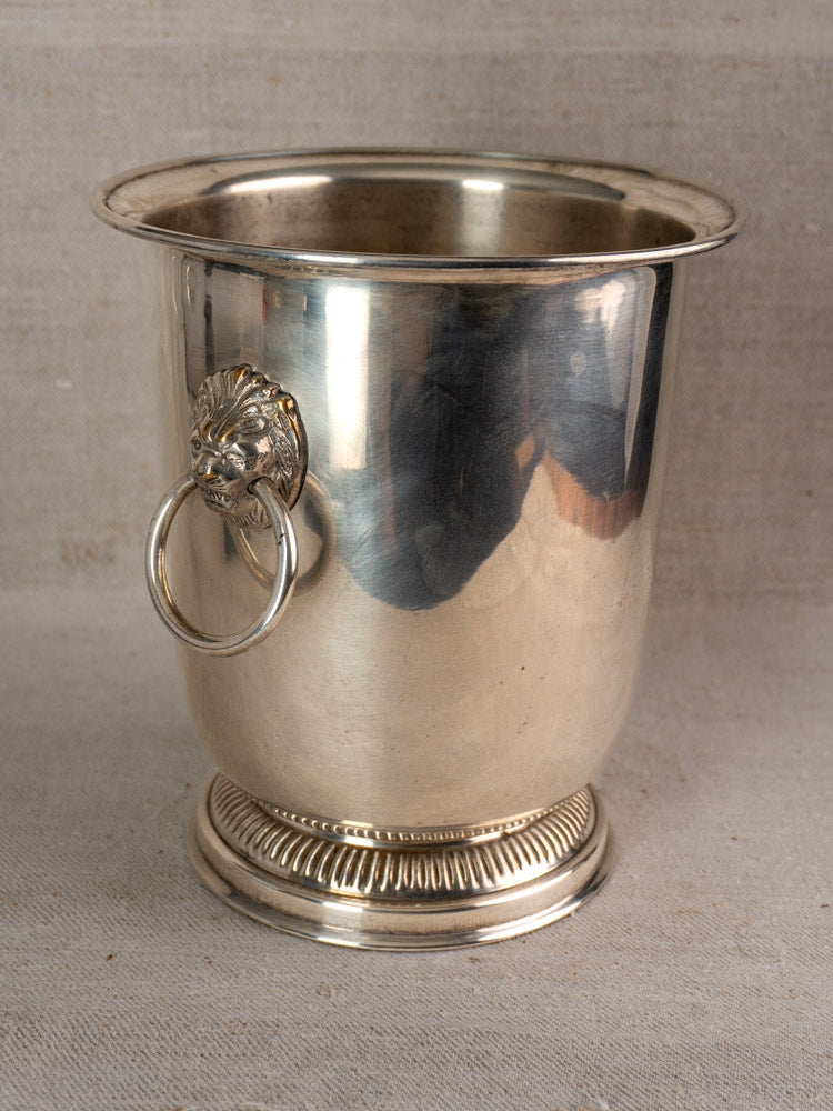 Vintage French silver-plated champagne bucket with lion head handles and front ring, set against neutral fabric backdrop.