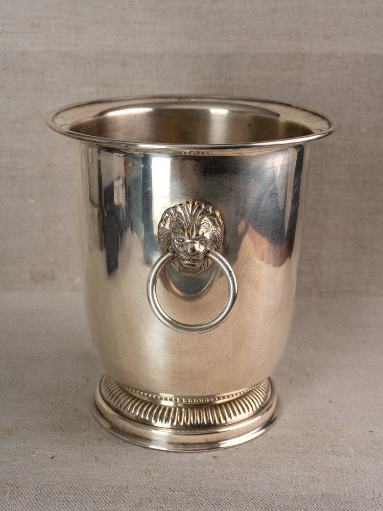 Vintage French silver-plated champagne bucket with lion head handles and front ring on a neutral fabric background.