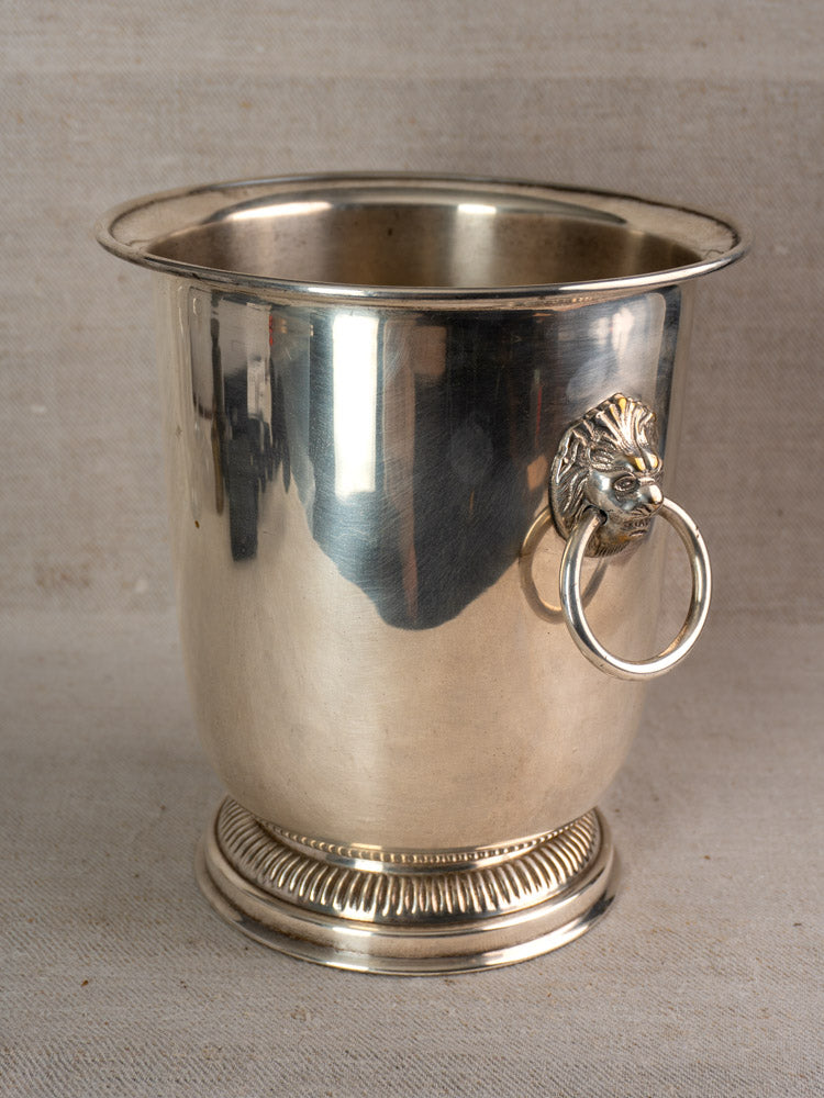 Vintage silver-plated French champagne bucket with lion head handles and textured base on neutral fabric backdrop.