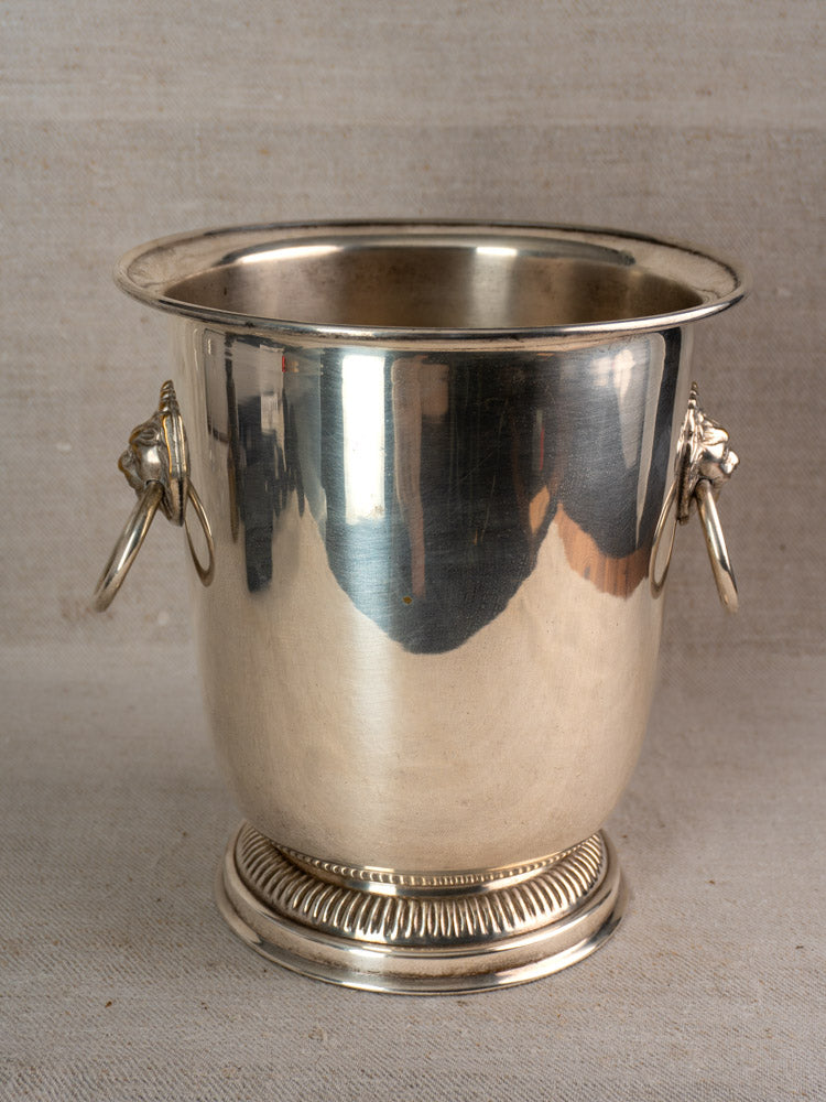 Vintage French silver-plated champagne bucket with lion head ring handles on a neutral fabric surface.