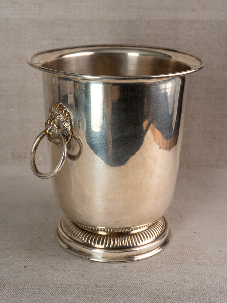 Silver-plated vintage French champagne bucket with lion head handles on a neutral fabric background.