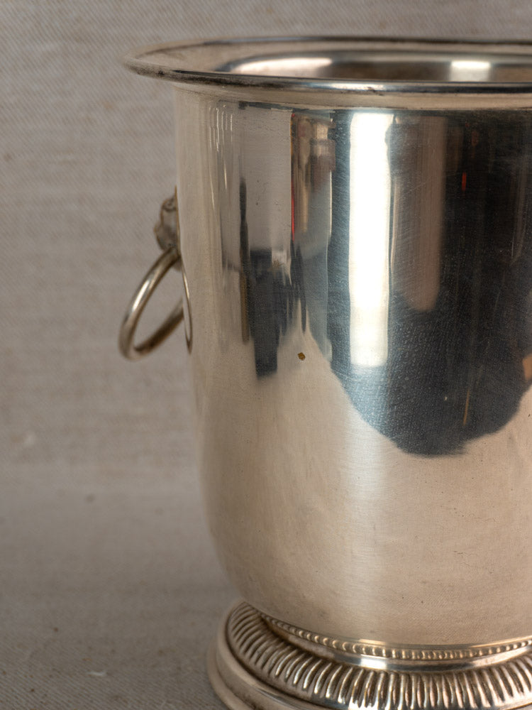 Vintage French silver plated champagne bucket with lion head handles and ornate base on a neutral textured background.