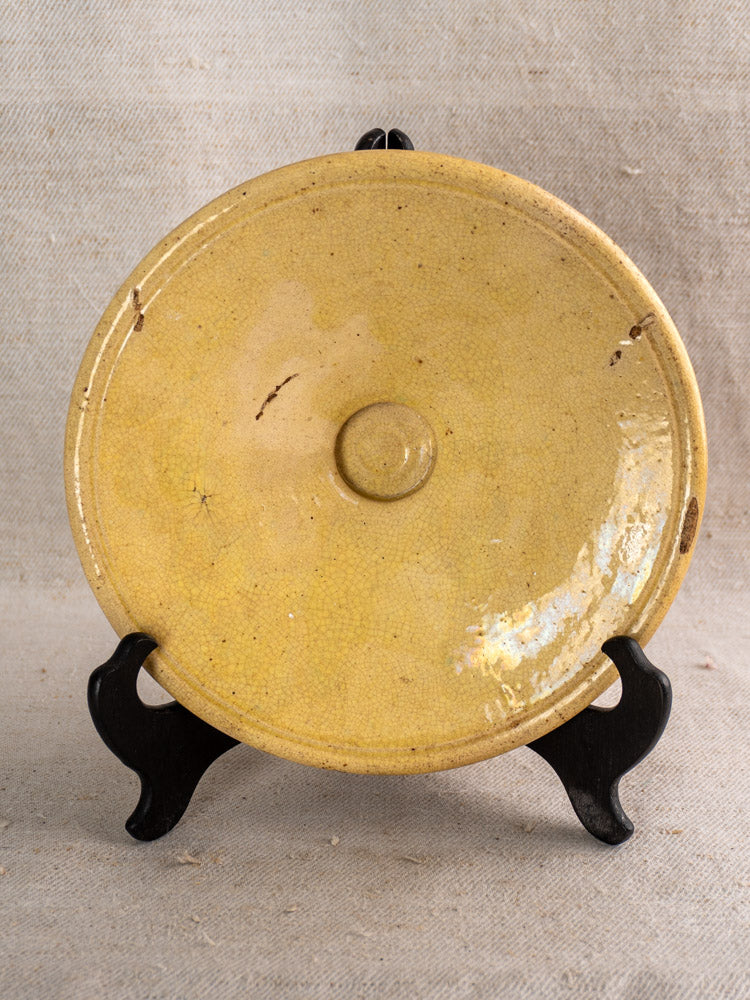 Antique French yellow ochre earthenware omelette plate on black stand with beige fabric backdrop, rustic dark marks visible.