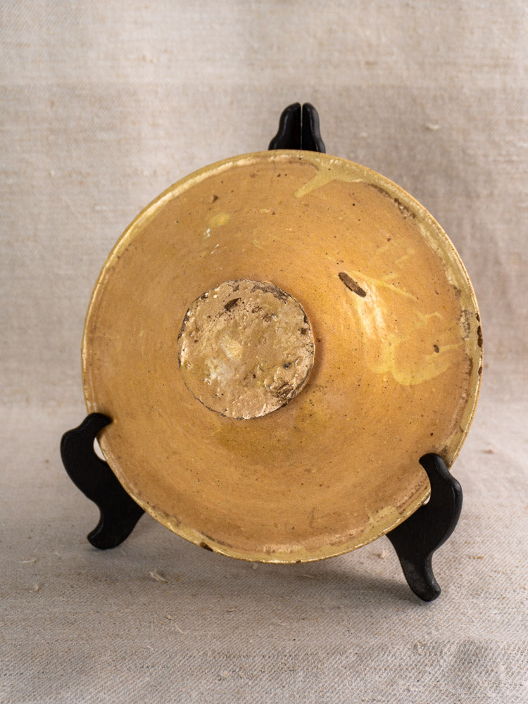 Antique French Provençal yellow ochre earthenware omelette plate on black stand, rough unglazed base shown, beige fabric backdrop.