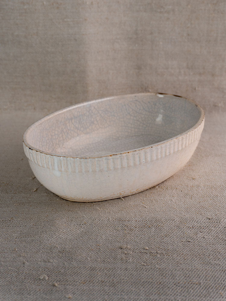 Antique French gratin dish, early 20th-century glazed earthenware with crackled white finish, on textured beige fabric.