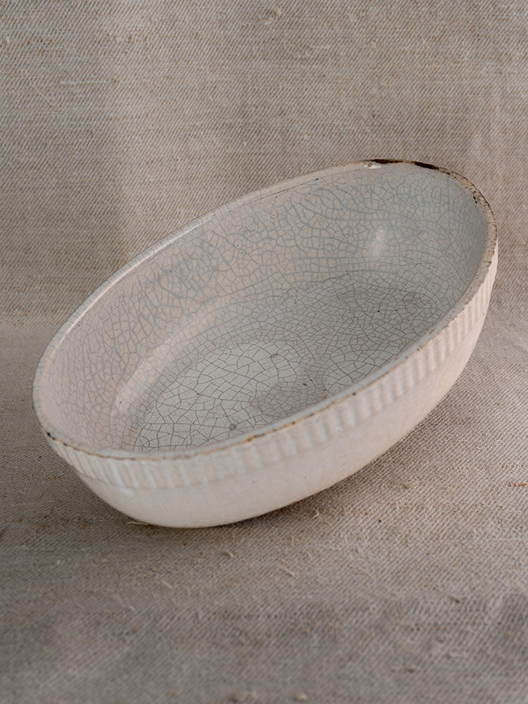 Antique French white glazed earthenware gratin dish with crackled finish and ribbed detail on textured beige fabric.