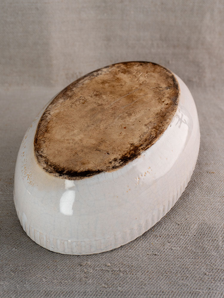 Antique French white glazed earthenware gratin dish, base exposed, on textured beige fabric, displaying patina and age.