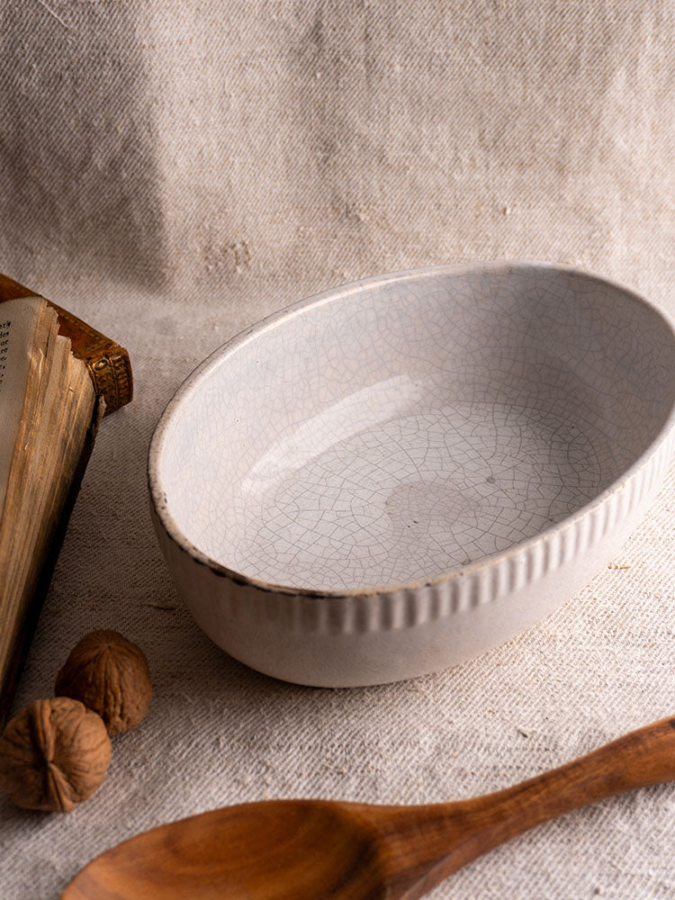 Antique French oval white earthenware gratin dish with crackle glaze on beige fabric with wooden spoon and walnuts.