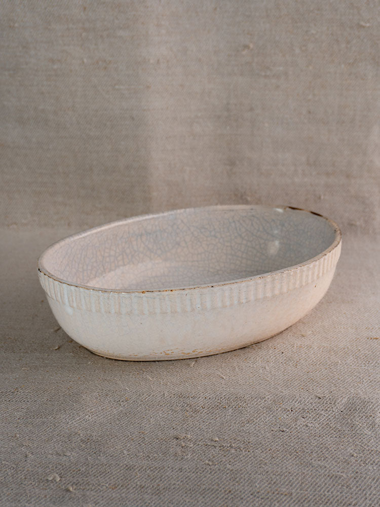 Antique French glazed earthenware gratin dish with crackled white finish and ridged sides on beige fabric.