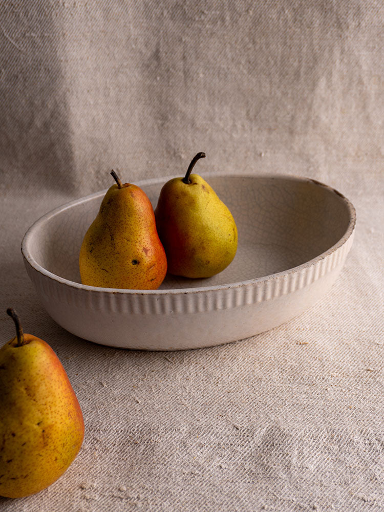 Antique French glazed earthenware gratin dish with two yellow pears, one pear on textured beige fabric, softly lit.