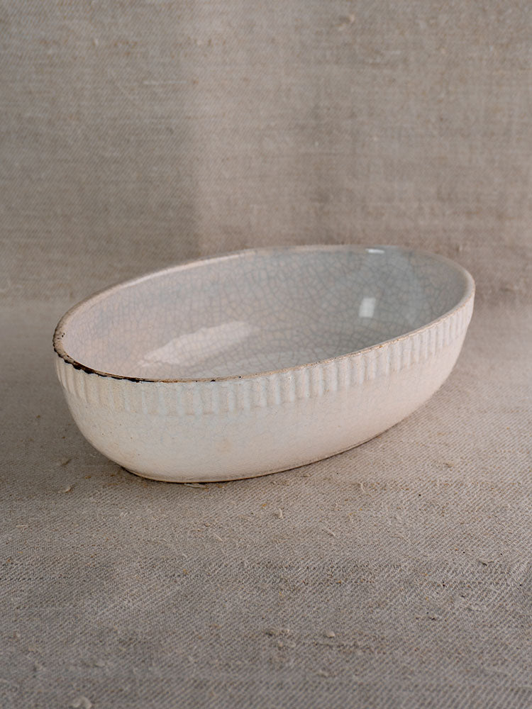 Antique French glazed earthenware gratin dish with textured rim and crackled off-white finish on beige woven fabric.