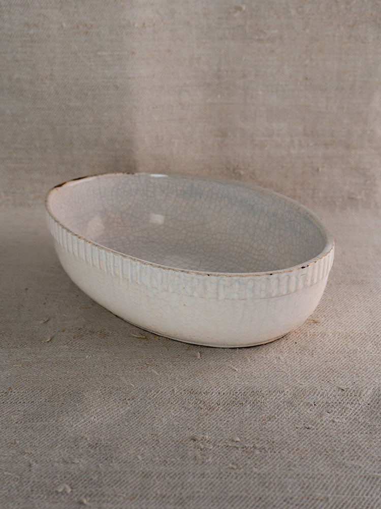 Antique French white glazed earthenware gratin dish with textured rim and crackle finish on neutral fabric background.