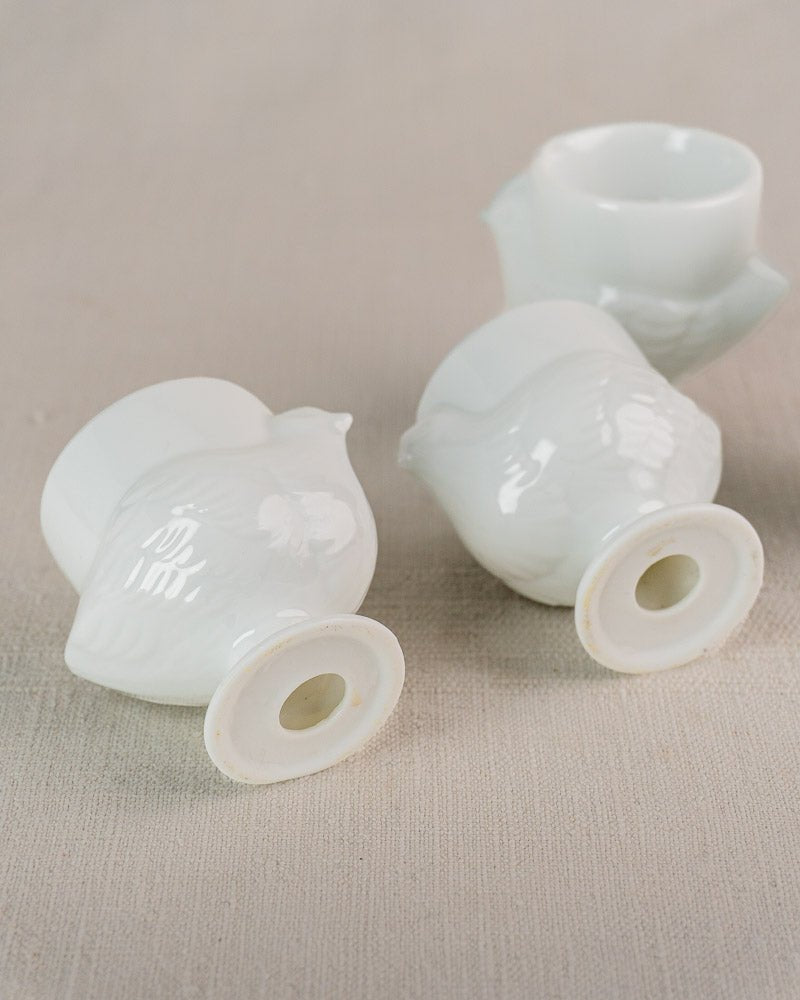 Four vintage French opaline glass egg cups shaped as hens, displayed on light beige fabric.